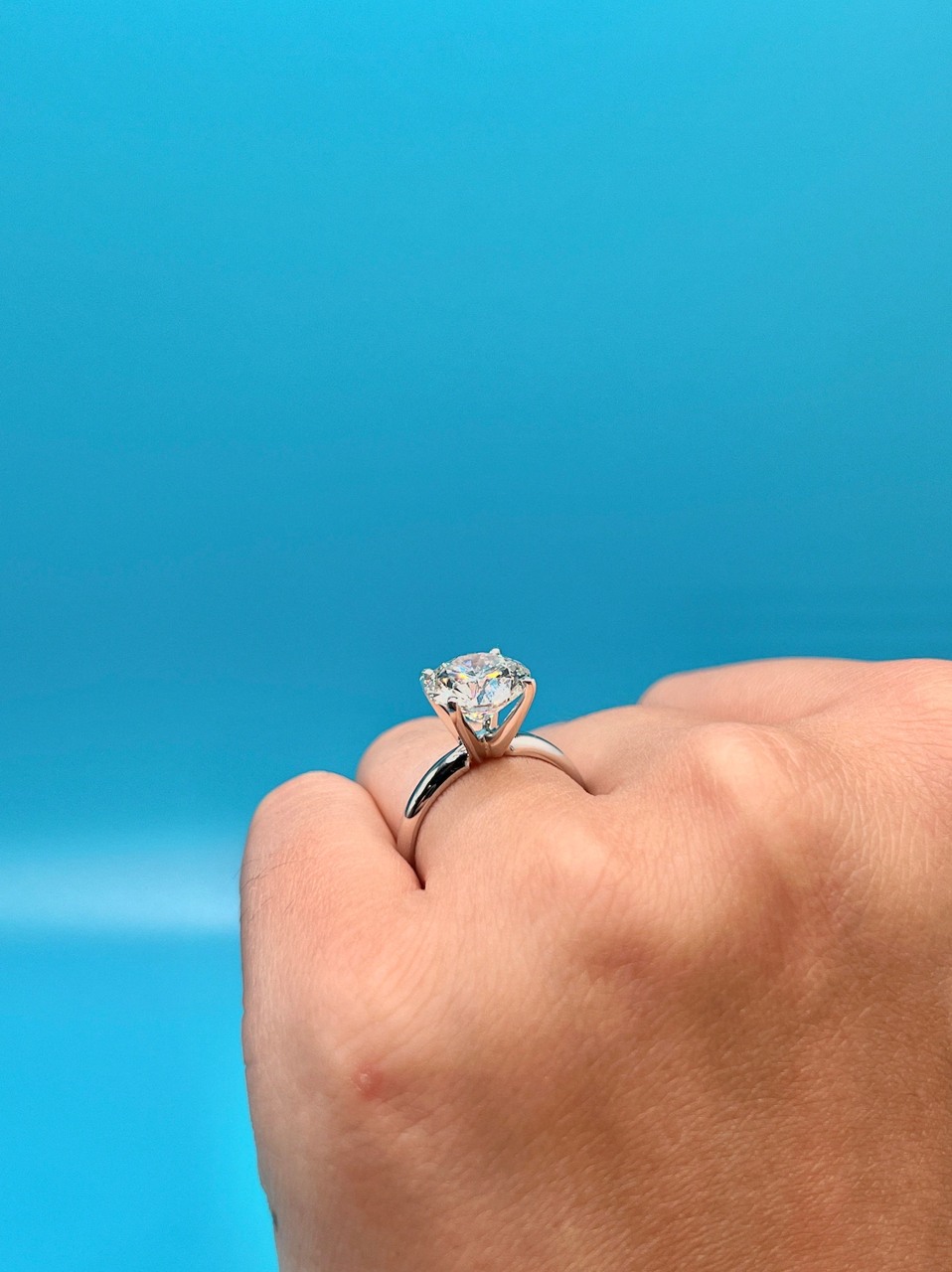 A close-up of a person's hand wearing a diamond engagement ring against a bright blue background.