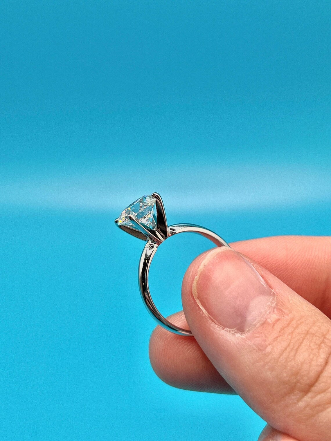 A hand holding a silver ring with a large diamond set in the center against a bright blue background.