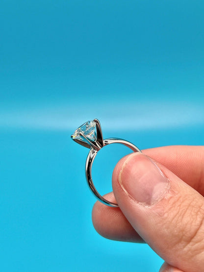 A hand holding a silver ring with a large diamond set in the center against a bright blue background.
