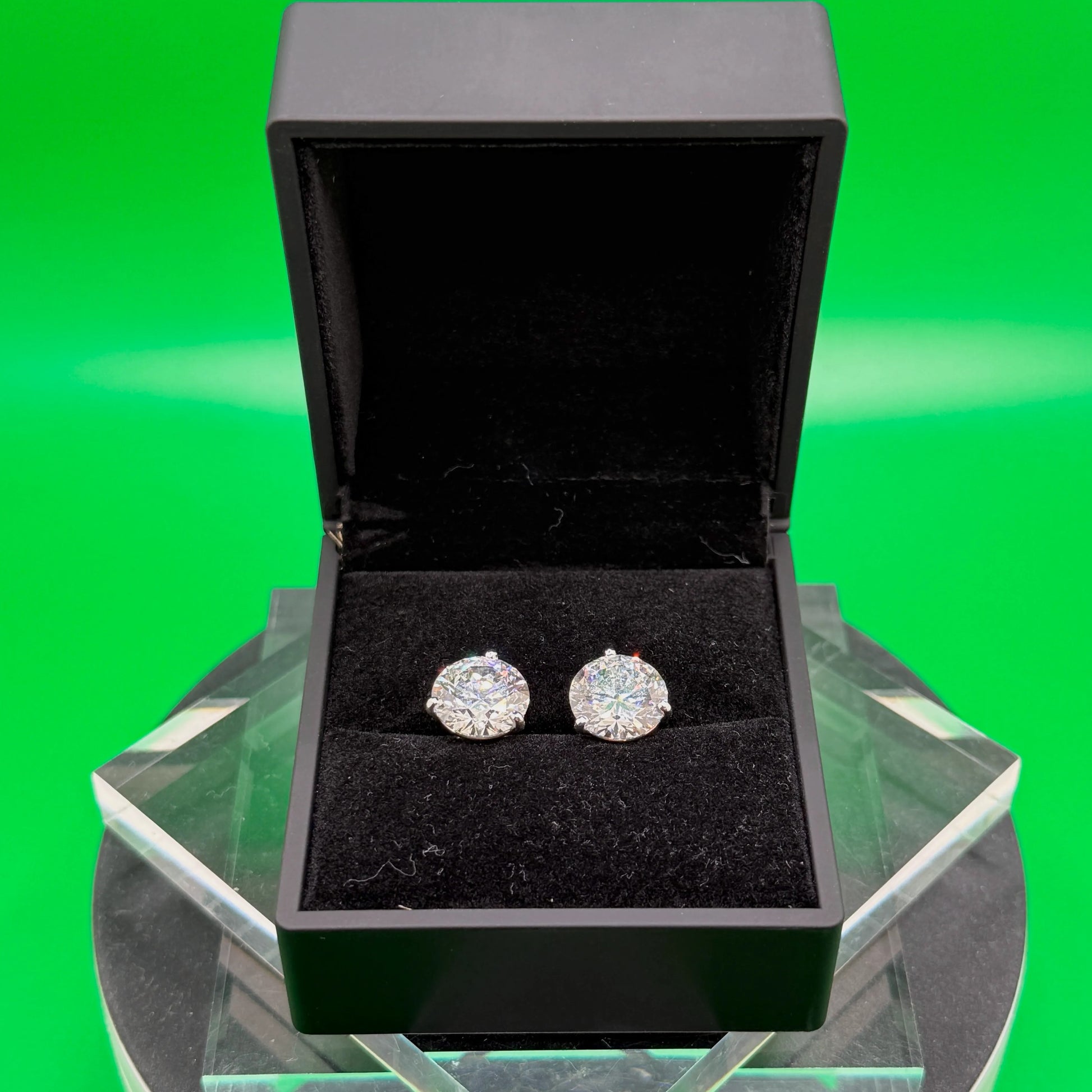 A pair of diamond earrings is displayed in a black jewelry box on a white pedestal against a green background.