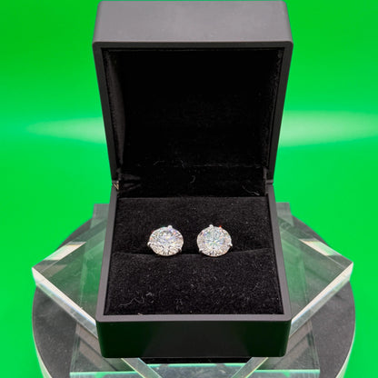 A pair of diamond earrings is displayed in a black jewelry box on a white pedestal against a green background.