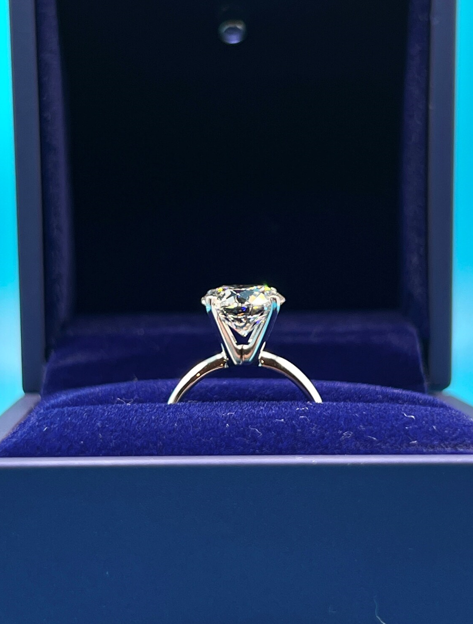 A diamond engagement ring is displayed in a blue velvet box against a dark background.