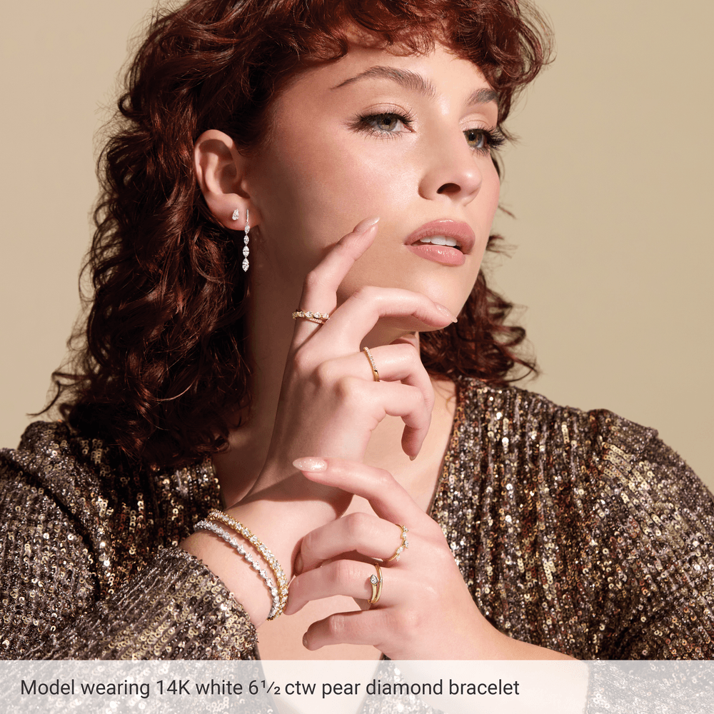 A woman with curly red hair wearing a sparkling gold sequin dress and a bracelet with pear diamonds, posing with her hand on her chin.