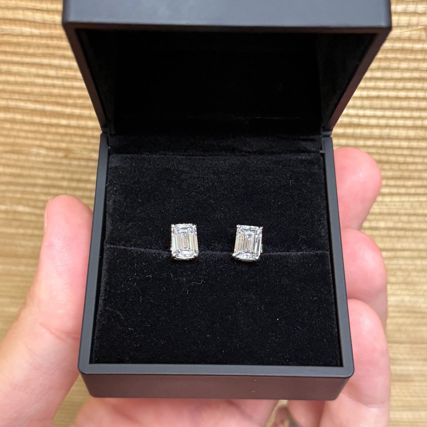 A person's hand holding a black jewelry box containing two diamond earrings.