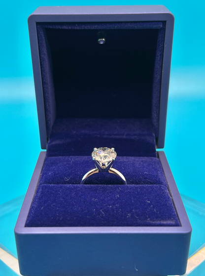 A diamond engagement ring is displayed in a blue velvet jewelry box against a teal background.