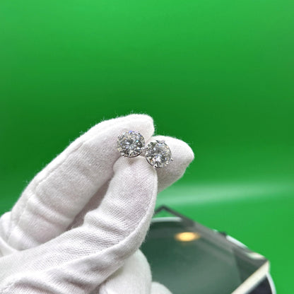 A hand wearing white gloves holds a pair of diamond earrings against a green background.