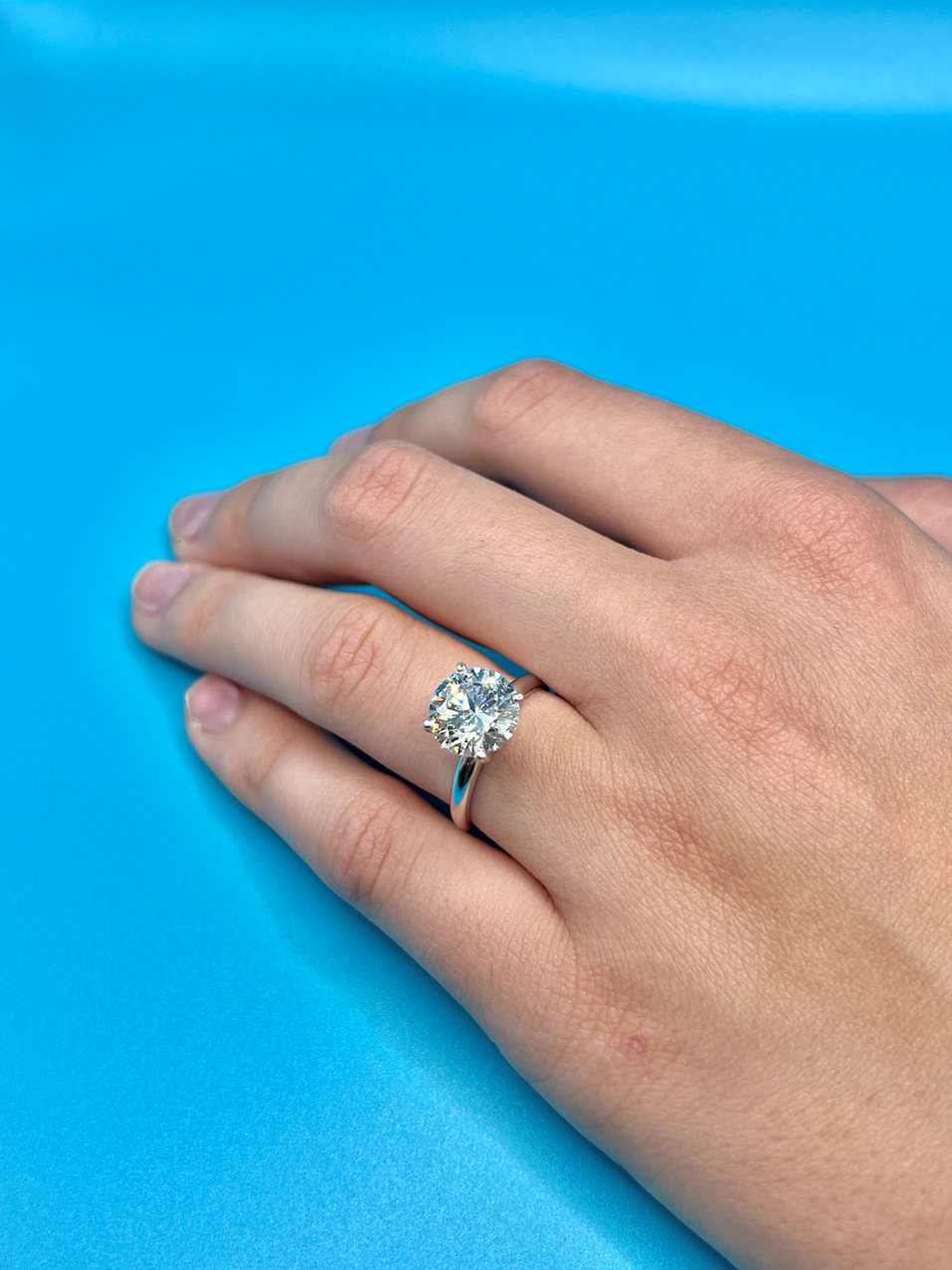 A close-up of a hand wearing a large diamond engagement ring against a bright blue background.