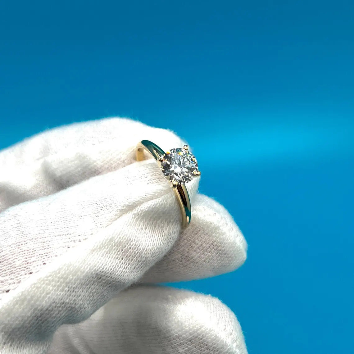 A close-up image of a person's hand wearing a gold ring with a diamond, against a blue background.