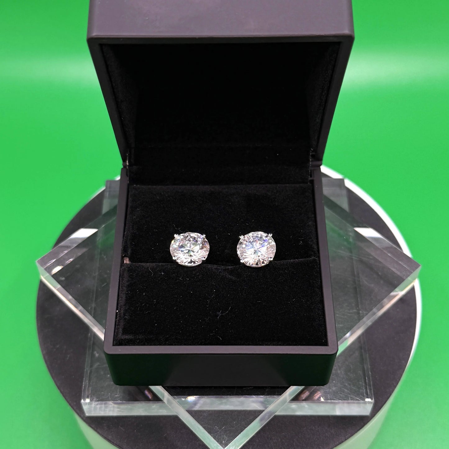 A pair of diamond earrings is displayed in a black jewelry box on a black stand, set against a green background.