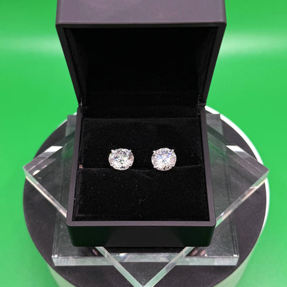 A pair of diamond earrings is displayed in a black jewelry box on a black stand, set against a green background.