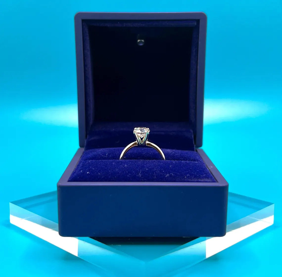 A diamond engagement ring is displayed in a blue velvet box against a blue background.