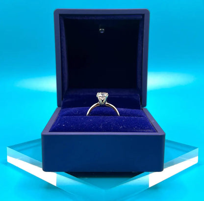 A diamond engagement ring is displayed in a blue velvet box against a blue background.