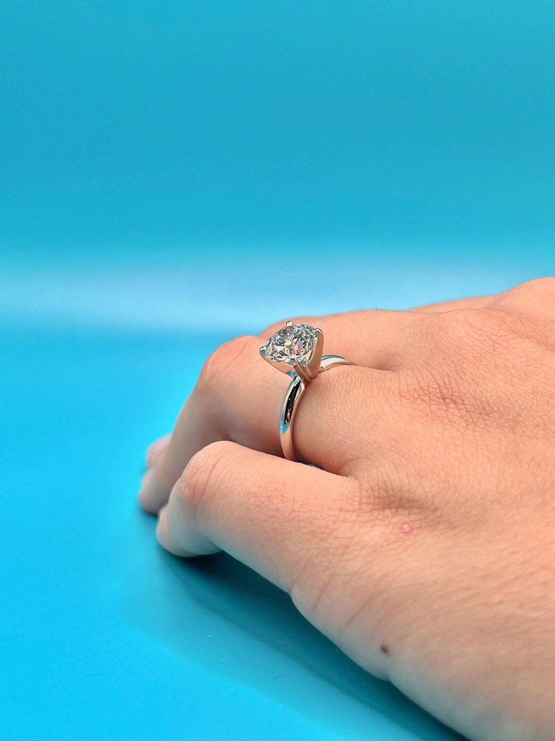 A close-up of a person's hand wearing a diamond engagement ring against a bright blue background.