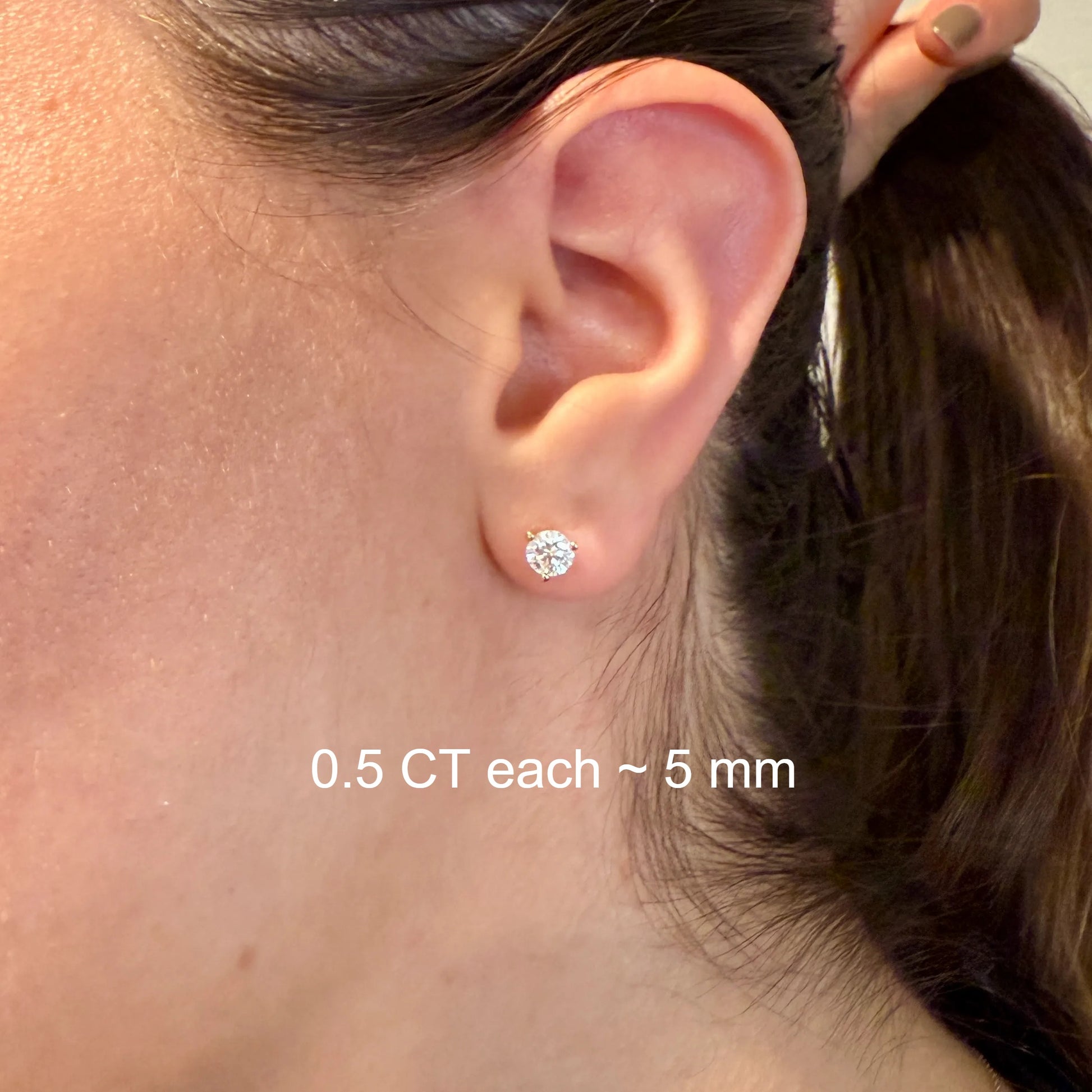 A close-up view of a person's ear with two diamond earrings, one larger and one smaller, both featuring a single diamond.