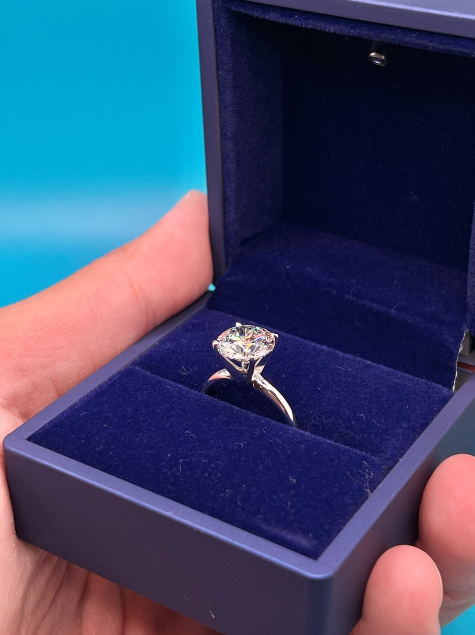 A hand holding a blue jewelry box containing a diamond engagement ring.