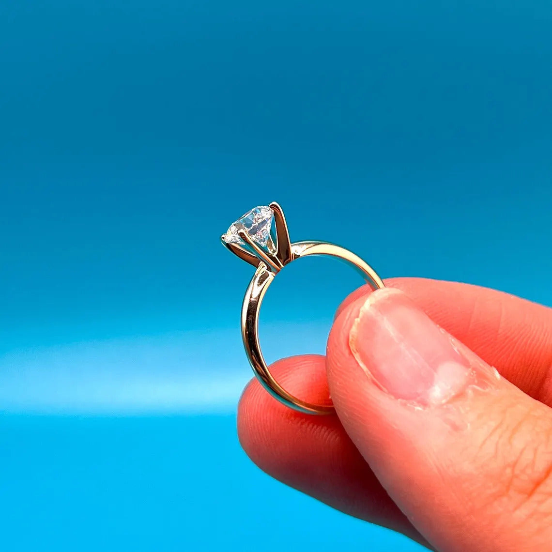 A close-up image of a person's hand holding a gold engagement ring with a diamond set in the center against a bright blue background.