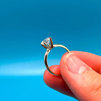 A close-up image of a person's hand holding a gold engagement ring with a diamond set in the center against a bright blue background.