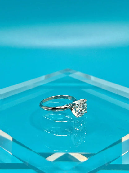 A diamond engagement ring with a large central diamond set in a silver band is displayed on a clear glass stand against a bright blue background.