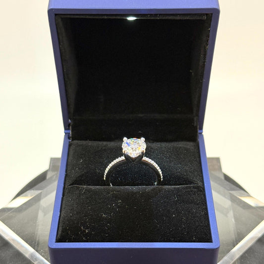 May include: A heart-shaped diamond ring displayed in a blue jewelry box. The ring features a large, clear diamond as the centerpiece, set on a delicate band. The band appears to be made of silver or white gold, with small accent stones. The ring is presented against a black velvet interior.