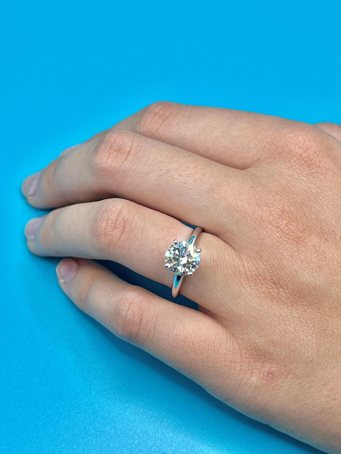 A close-up of a hand wearing a diamond engagement ring against a blue background.