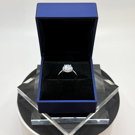 May include: A diamond ring in a blue jewelry box. The ring features a central, round-cut diamond surrounded by a halo of smaller diamonds. The ring sits on a black velvet interior. The jewelry box is a deep blue color and is open, displaying the ring.