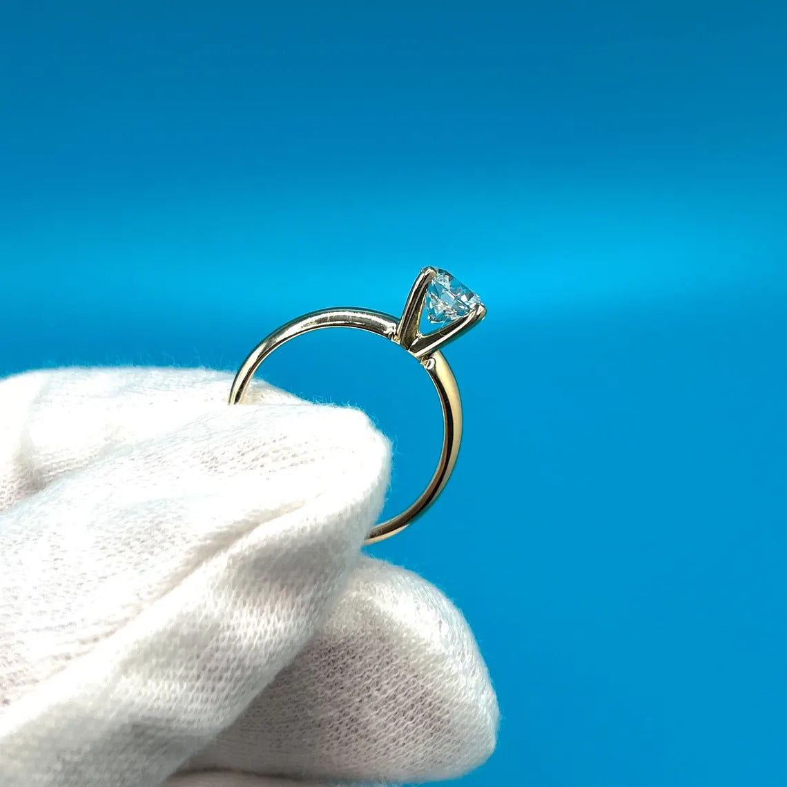 A close-up image of a gold ring with a diamond set in the center, being held in a gloved hand against a bright blue background.