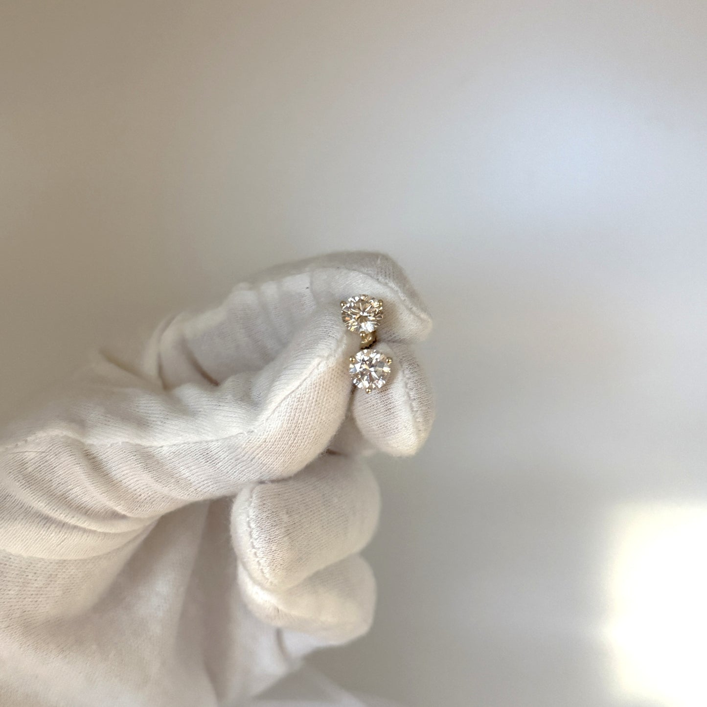 A hand wearing a white glove holds a pair of diamond earrings against a blurred background.