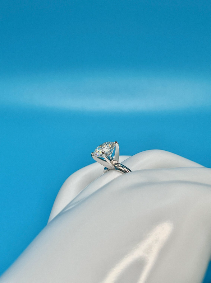 A close-up view of a diamond engagement ring on a white mannequin hand against a blue background.