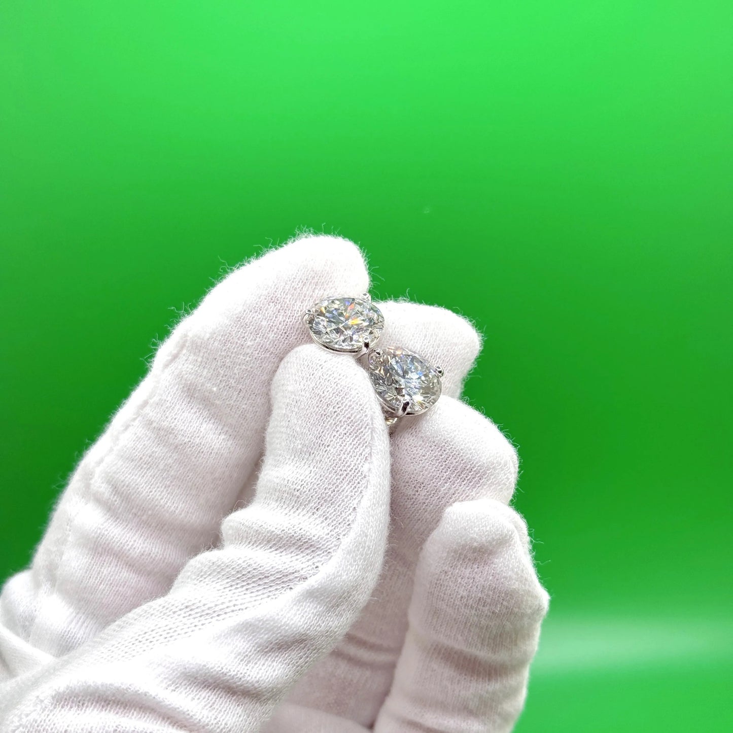 A hand wearing white gloves holds a pair of diamond earrings against a green background.